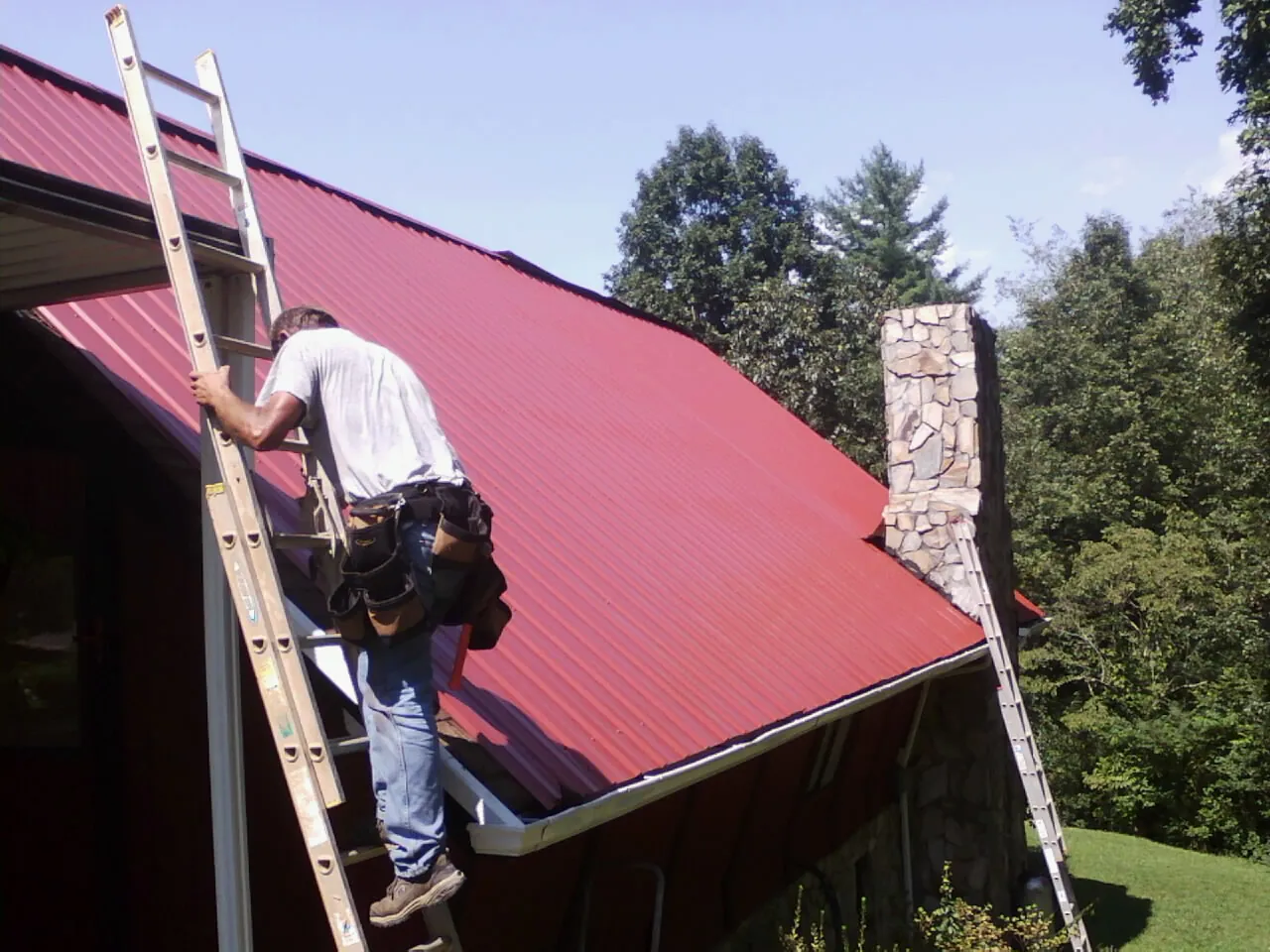 Roofer on a ladder beside a red metal roof and stone chimney — Soffit & Fascia Repair by Roofing Brothers in South Portland